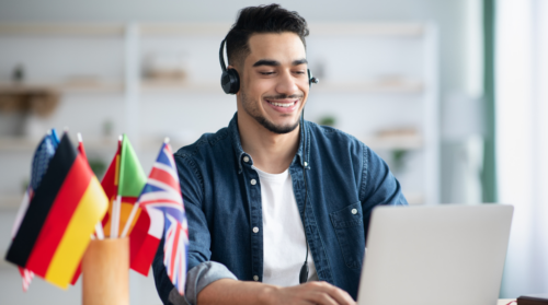 Na imagem, há um jovem branco, de cabelo preto e barba, blusa branca e camisa jeans, fones nos ouvidos, em frente a um notebook. Ao lado do rapaz, há um copo com bandeirinhas de diversos países. Ele está em, aparentemente, uma sala de aula, desfocada de fundo. Fim da descrição.