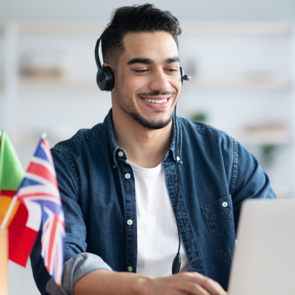 Na imagem, há um jovem branco, de cabelo preto e barba, blusa branca e camisa jeans, fones nos ouvidos, em frente a um notebook. Ao lado do rapaz, há um copo com bandeirinhas de diversos países. Ele está em, aparentemente, uma sala de aula, desfocada de fundo. Fim da descrição.